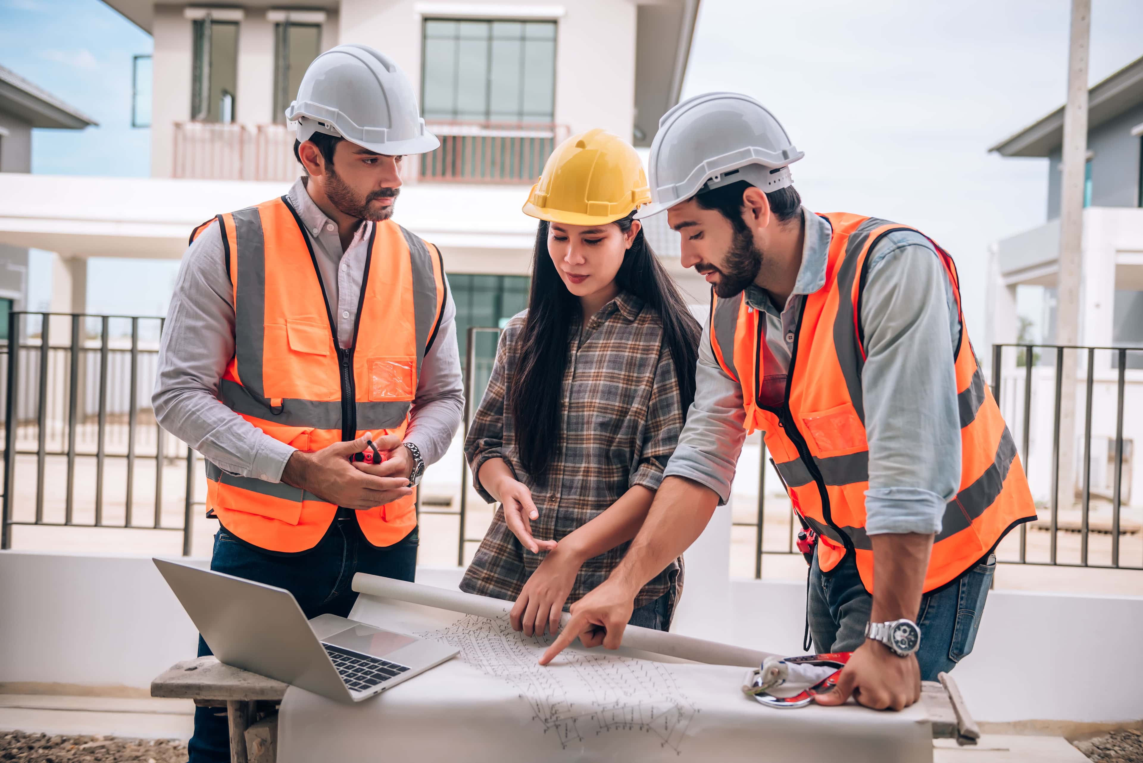 Construction project managers and engineers in safety gear reviewing blueprints outside a residential project